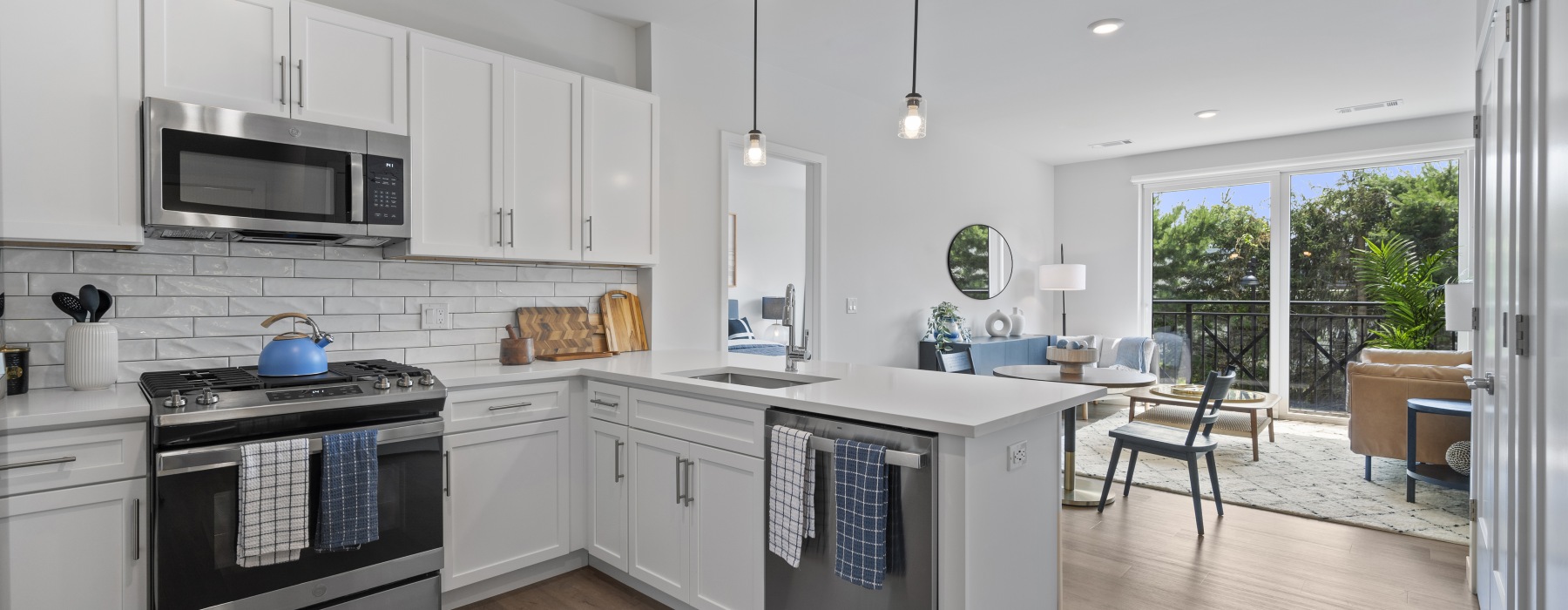 Model kitchen and living room at our apartments for rent in Wilton, CT featuring wood grain floor paneling and a view of lush greenery outside apartment